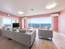 A modern living room with a large sofa, a smaller armchair, a flat-screen TV, and a large window offering a view of the sea.