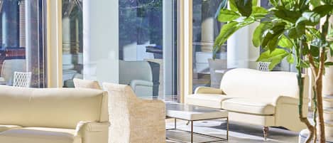 Una sala de estar moderna con un sofá beige, una silla de mimbre y un sillón blanco. Amplias ventanas ofrecen vistas al exterior.