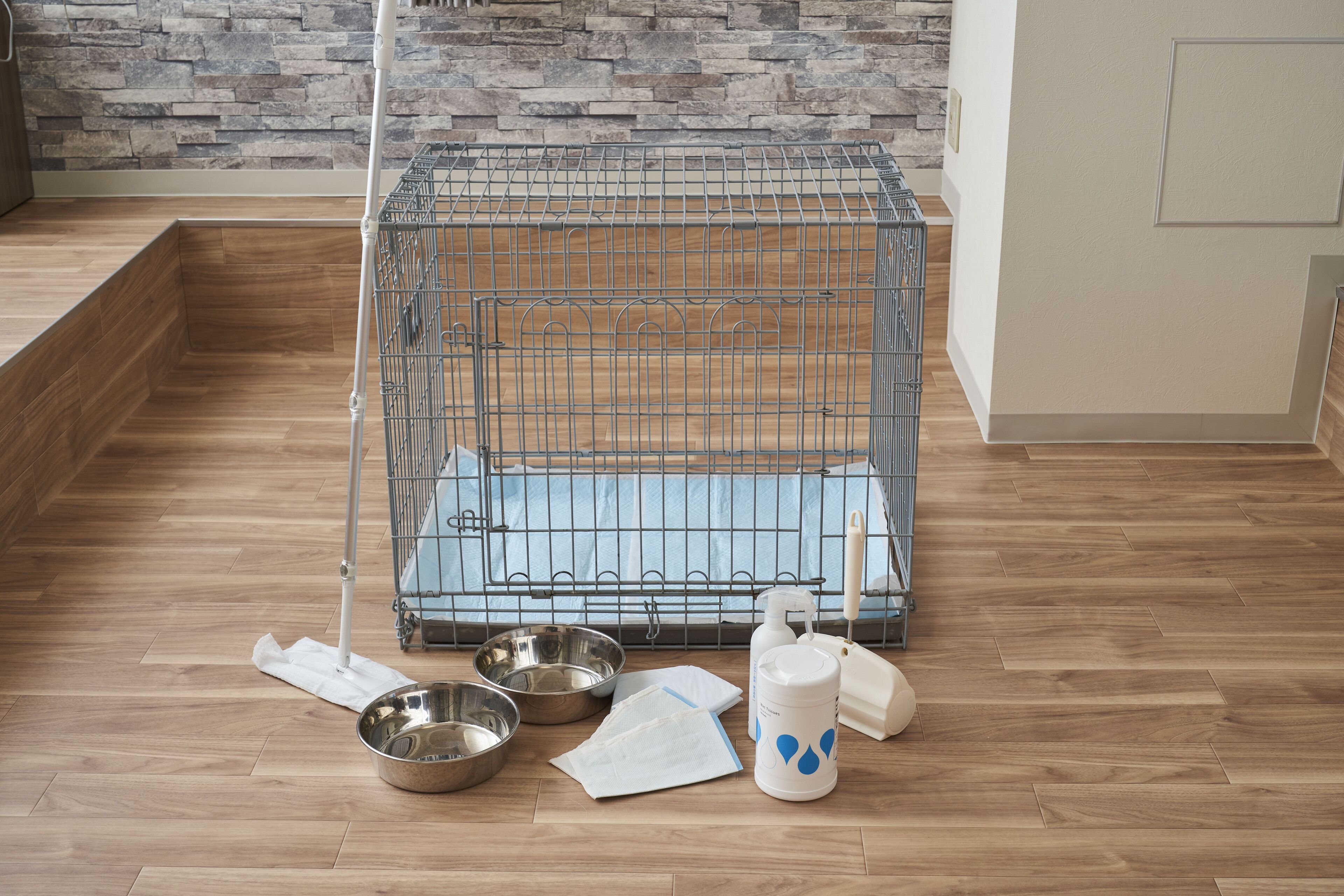 A pet cage with a wooden floor, metal food bowls, a white water dispenser, and a white brush.