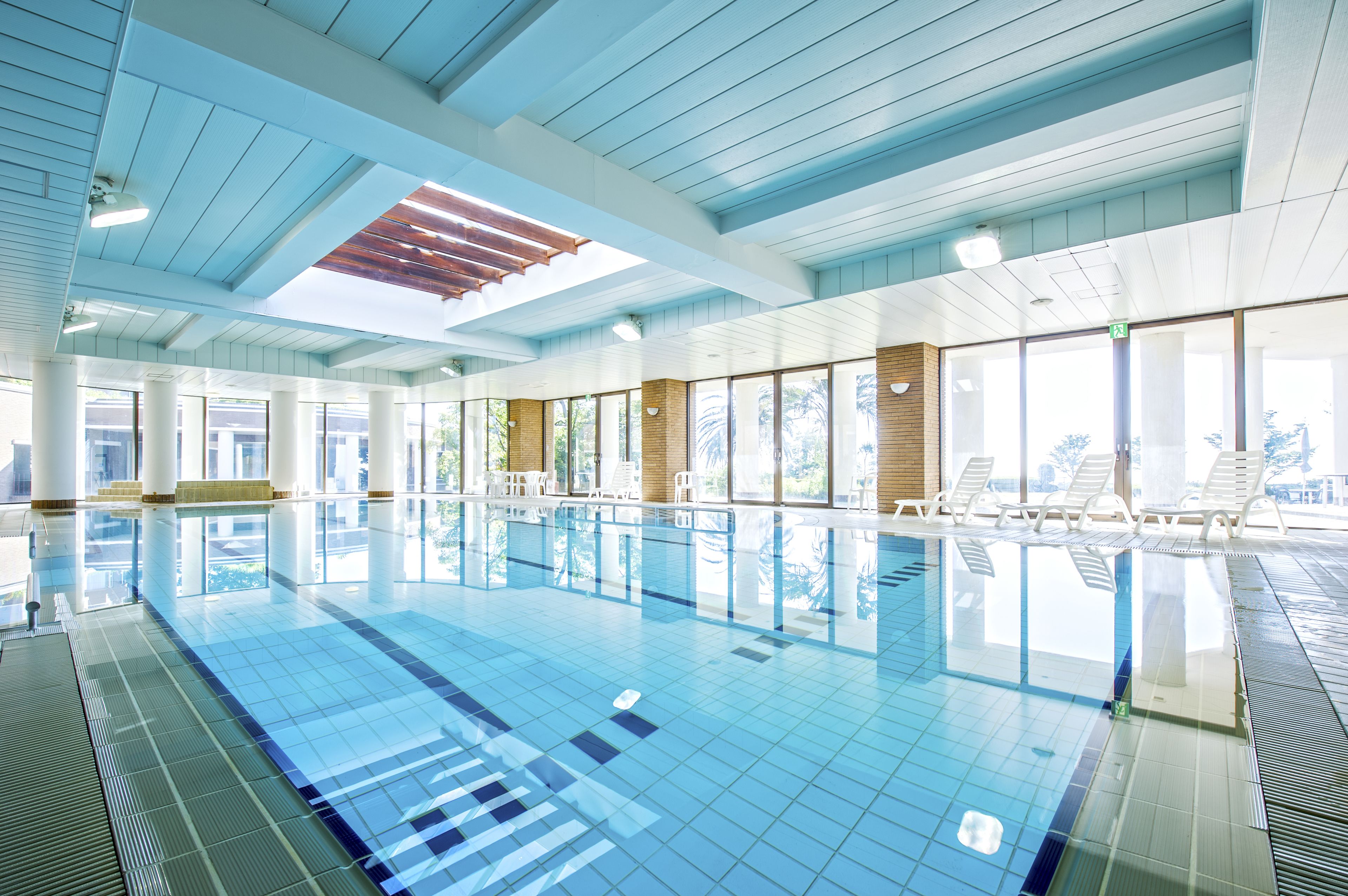A large indoor swimming pool with clear blue water, surrounded by white lounge chairs and a high ceiling with wooden beams.