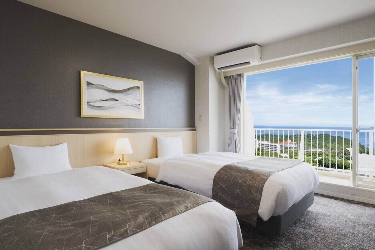 Une chambre d’hôtel avec deux lits, une table de chevet avec une lampe, un tableau encadré au mur et un balcon avec vue.