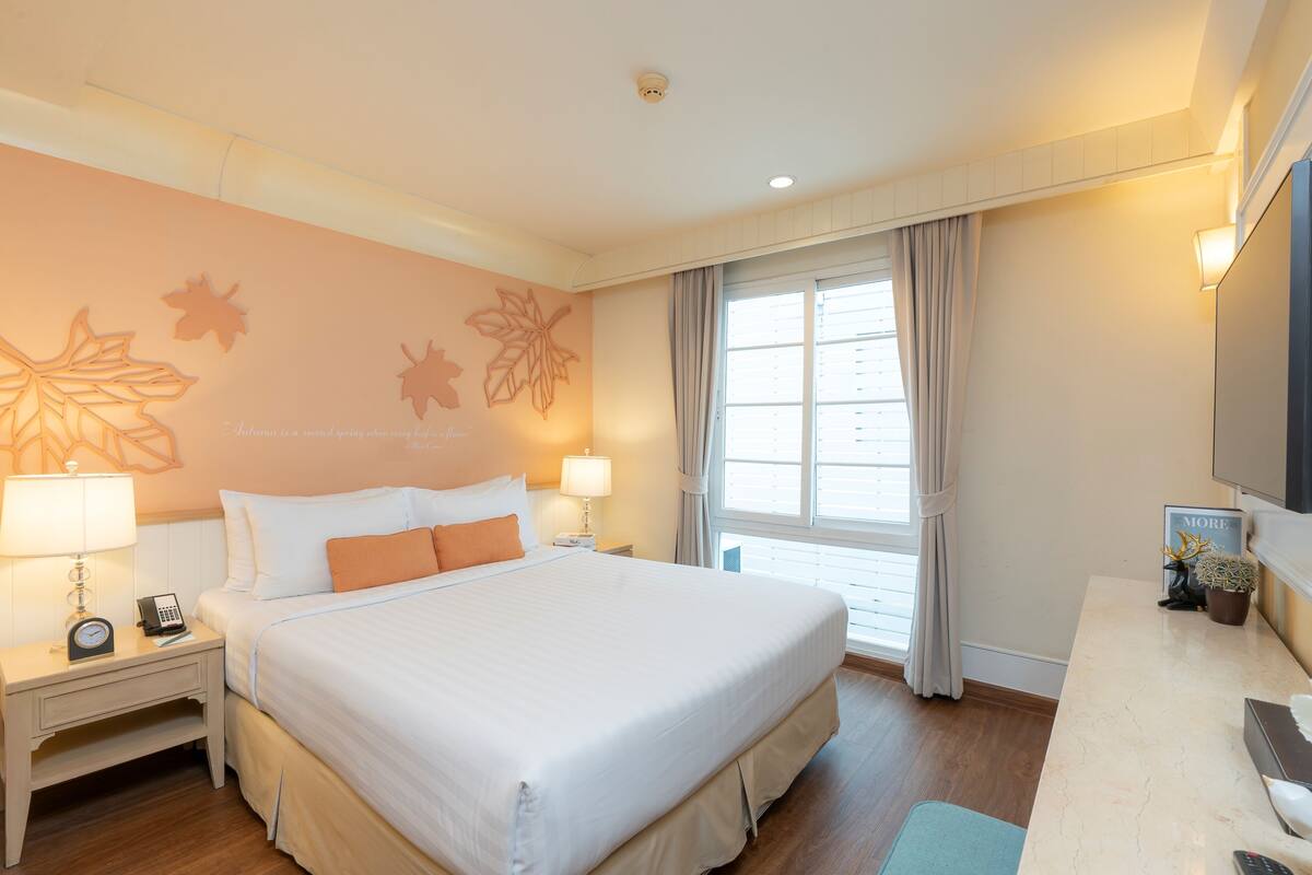 A hotel room with a large bed, bedside lamps, a desk with a chair, a window with curtains, and wall art.