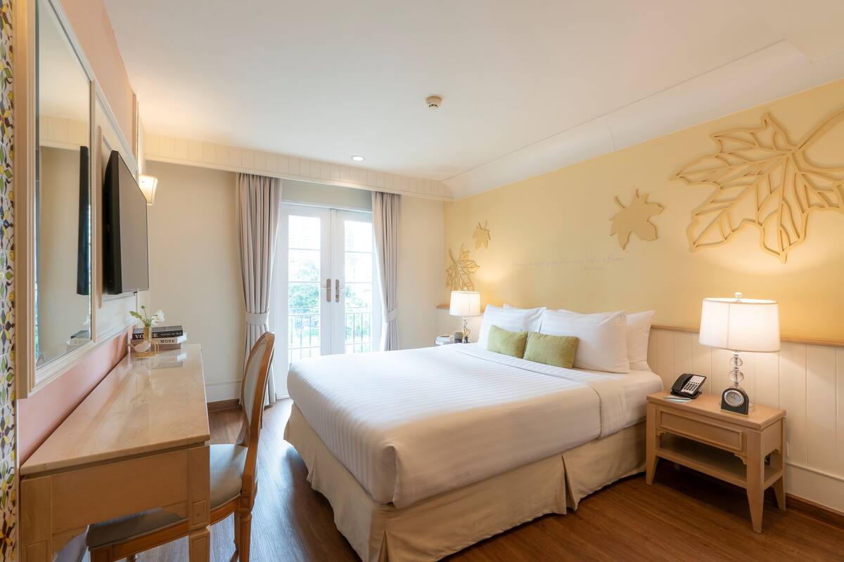 A hotel room with a bed, bedside table, lamp, chair, desk, and a large window with curtains.
