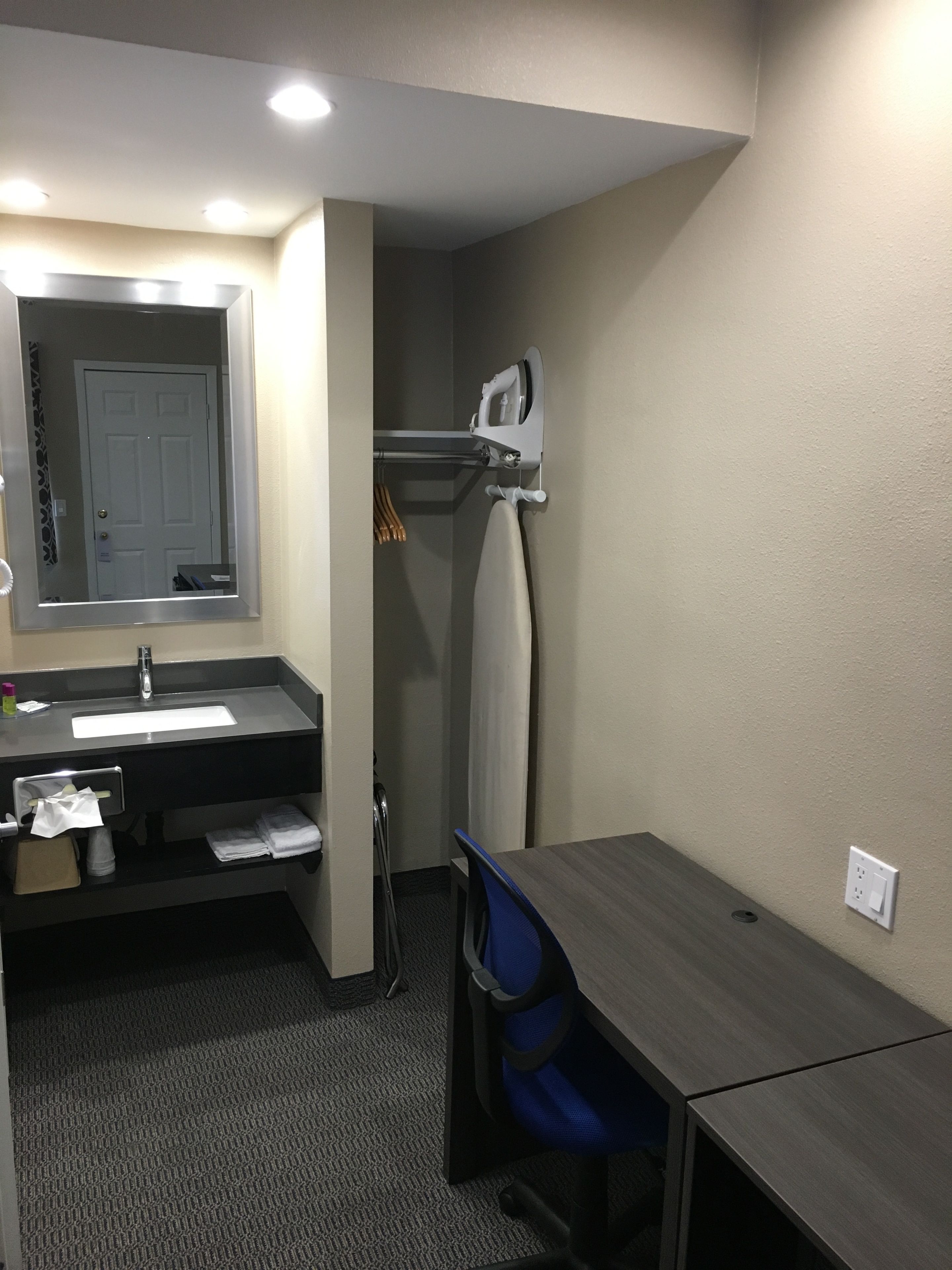 A hotel room with a desk, chair, sink, mirror, and a hanging towel rack.