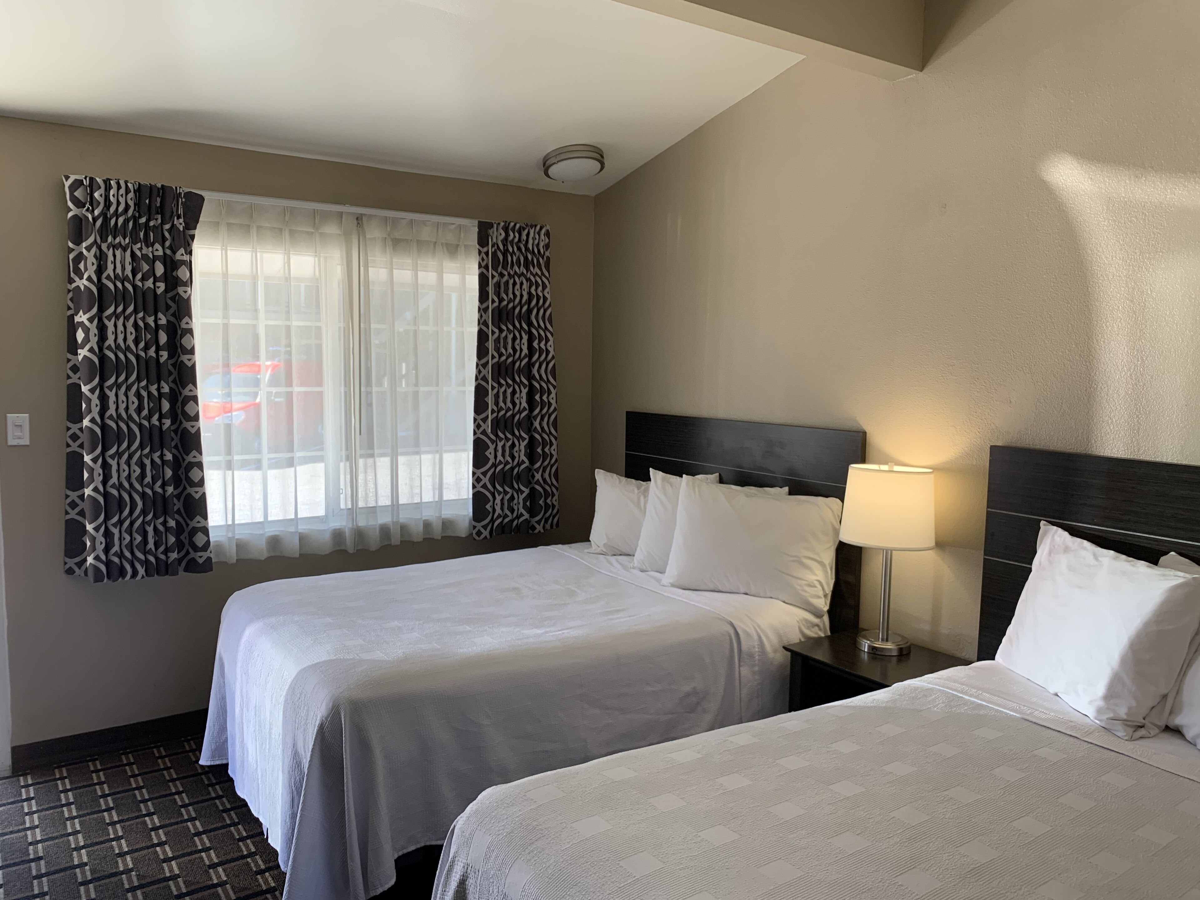 Quarto de hotel com duas camas, janela com cortinas estampadas, luminária de cabeceira e piso padronizado.