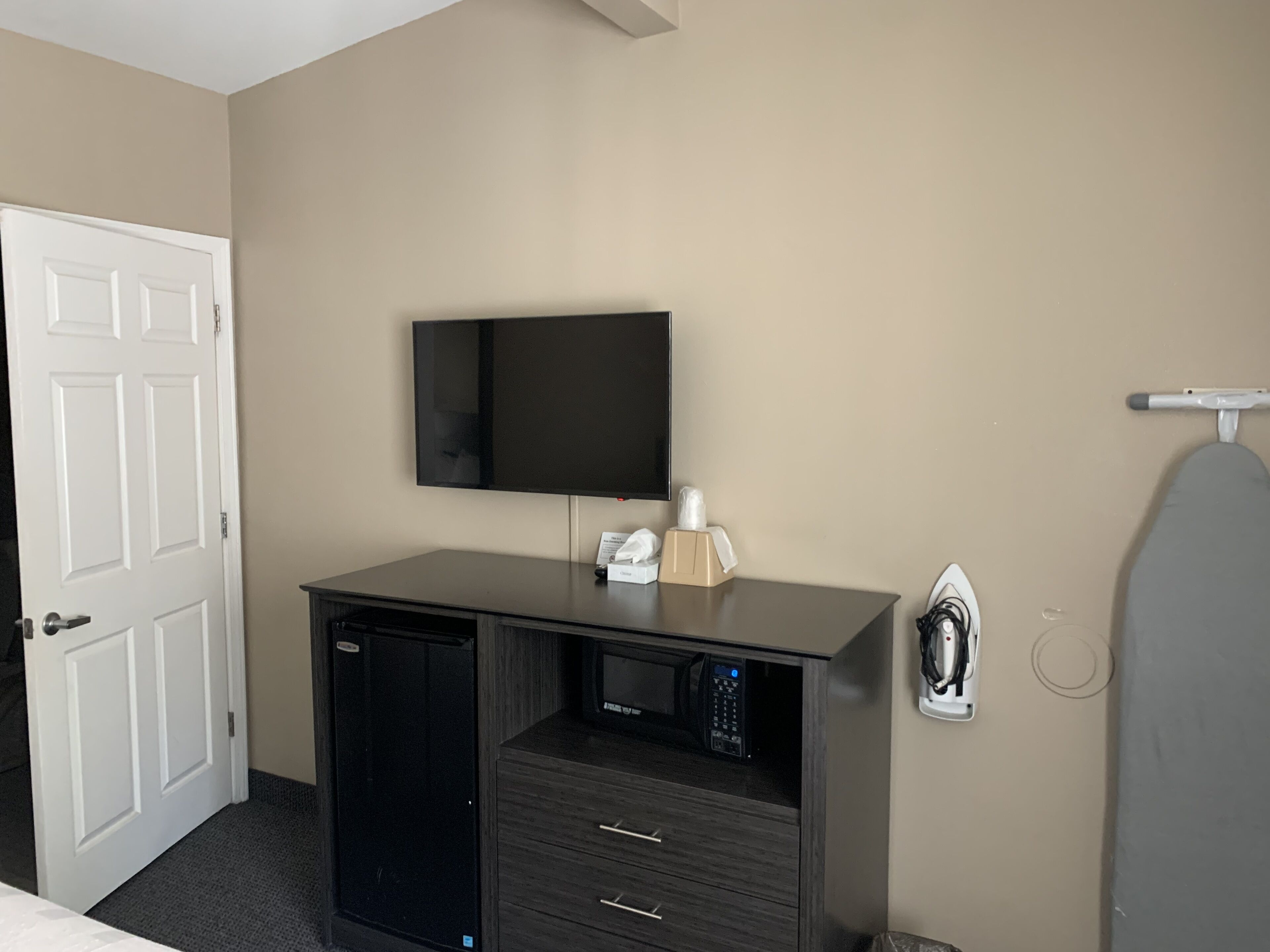 Hotelkamer met een flatscreen televisie aan de muur, een kleine koelkast, een klein bureau met lades en een wandplank met een strijkplank.