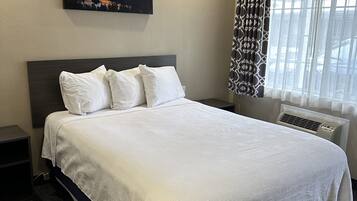 A hotel room with a large bed, a painting on the wall, a window with patterned curtains, and a wall-mounted air conditioner.