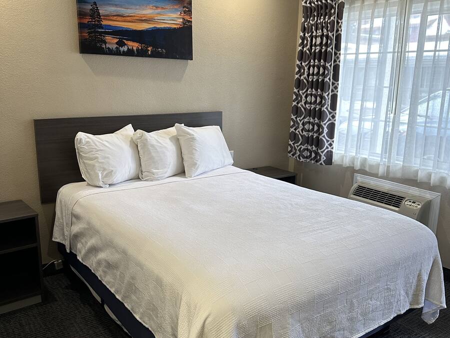 A hotel room with a large bed, a painting on the wall, a window with patterned curtains, and a wall-mounted air conditioner.