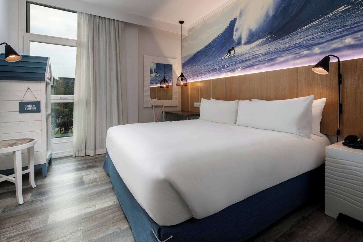 Habitació d'hotel amb un llit gran, una taula de treball i una cadira. Hi ha una finestra amb cortines, una paret amb un mural de surf i un cartell que diu "Gone Surfing".