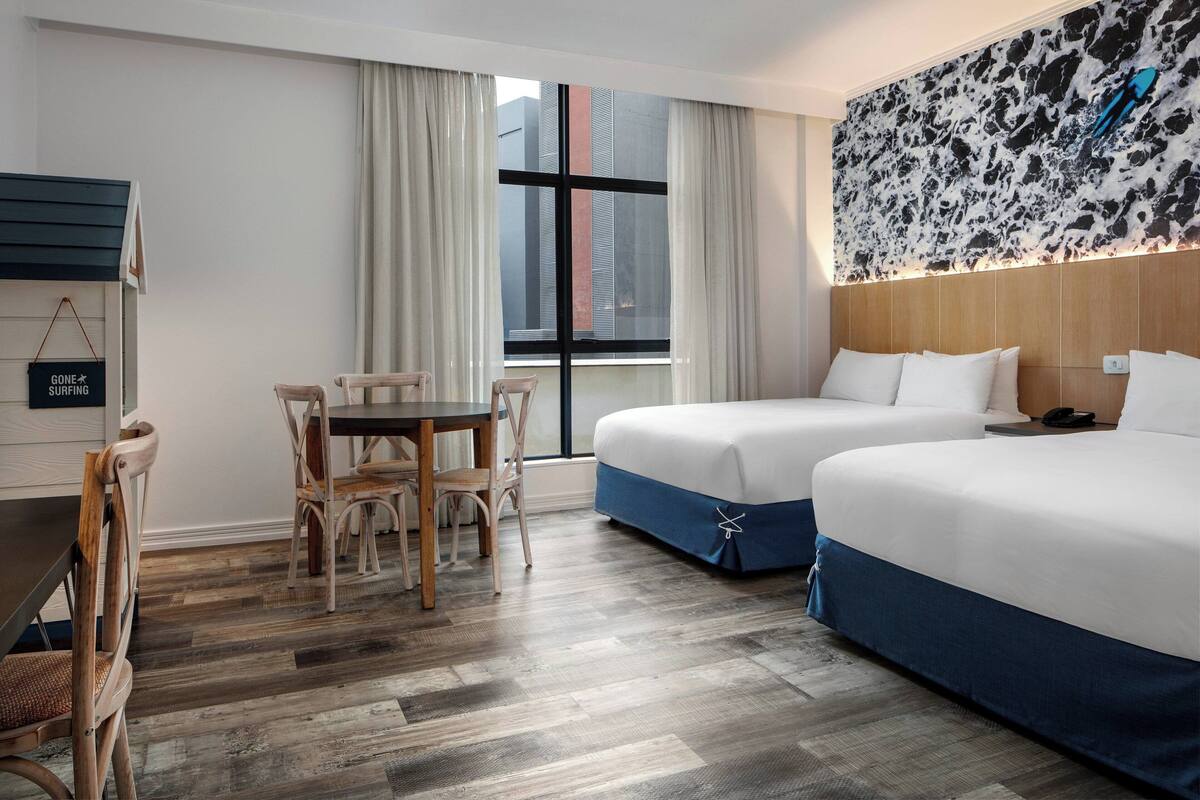 Habitació d'hotel amb dos llits, una taula de menjador amb cadires, una finestra gran amb cortines i una obra d'art penjada a la paret.