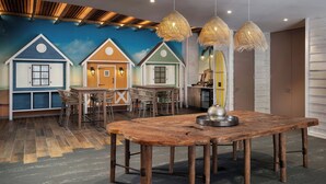 Uma área de refeições com mesa e cadeiras de madeira, um mural com casas na parede e luminárias de vime penduradas.