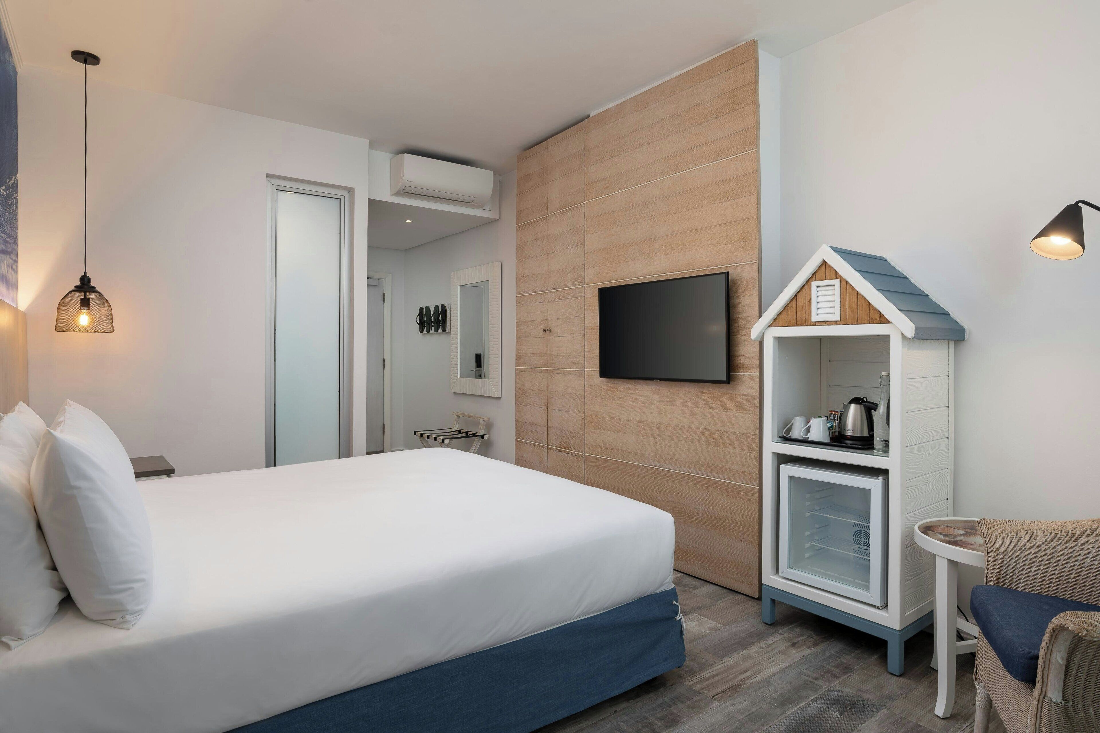 Una habitación de hotel con una cama, un televisor montado en la pared, un refrigerador pequeño y una silla de mimbre.