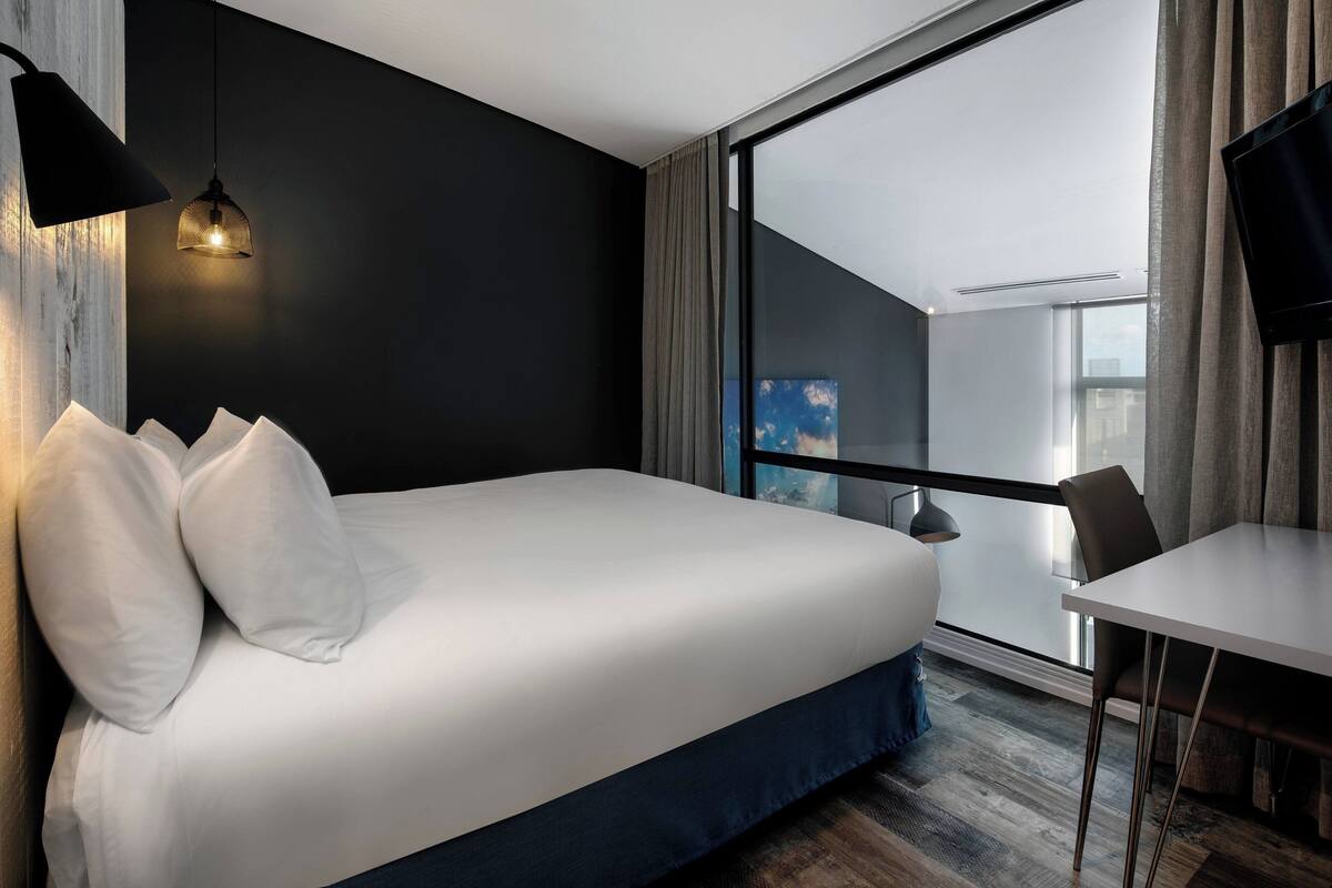 Habitació d'hotel moderna amb un llit gran, un escriptori i vista a la ciutat a través d'una finestra ampla.