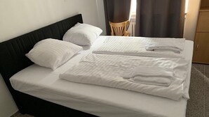 Un llit ben fet amb llençols i almoades blanques, un capçal fosc i una cadira de fusta al fons.
