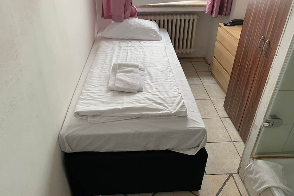 A small, neatly made bed with white linens and a folded towel, a wooden dresser, and a radiator under a window with pink curtains.