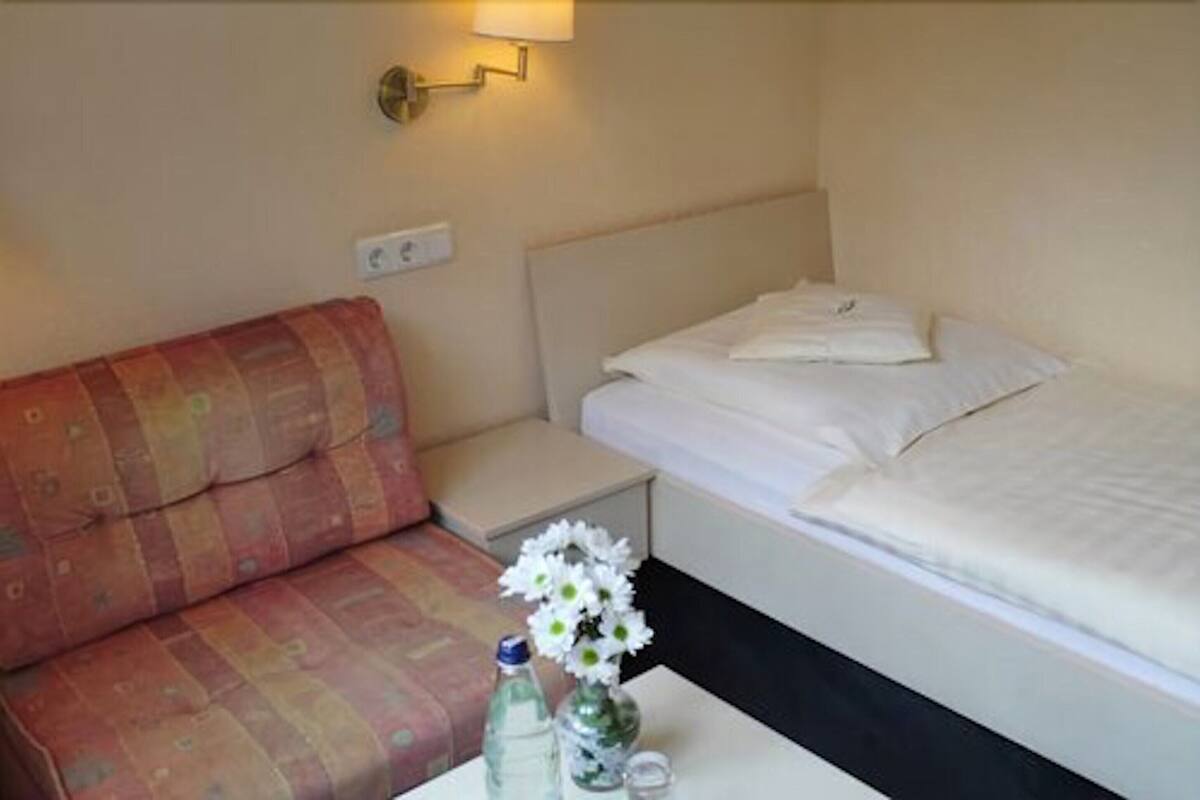 A hotel room with a bed, a small bedside table, a nightstand, a sofa, and a wall-mounted lamp.