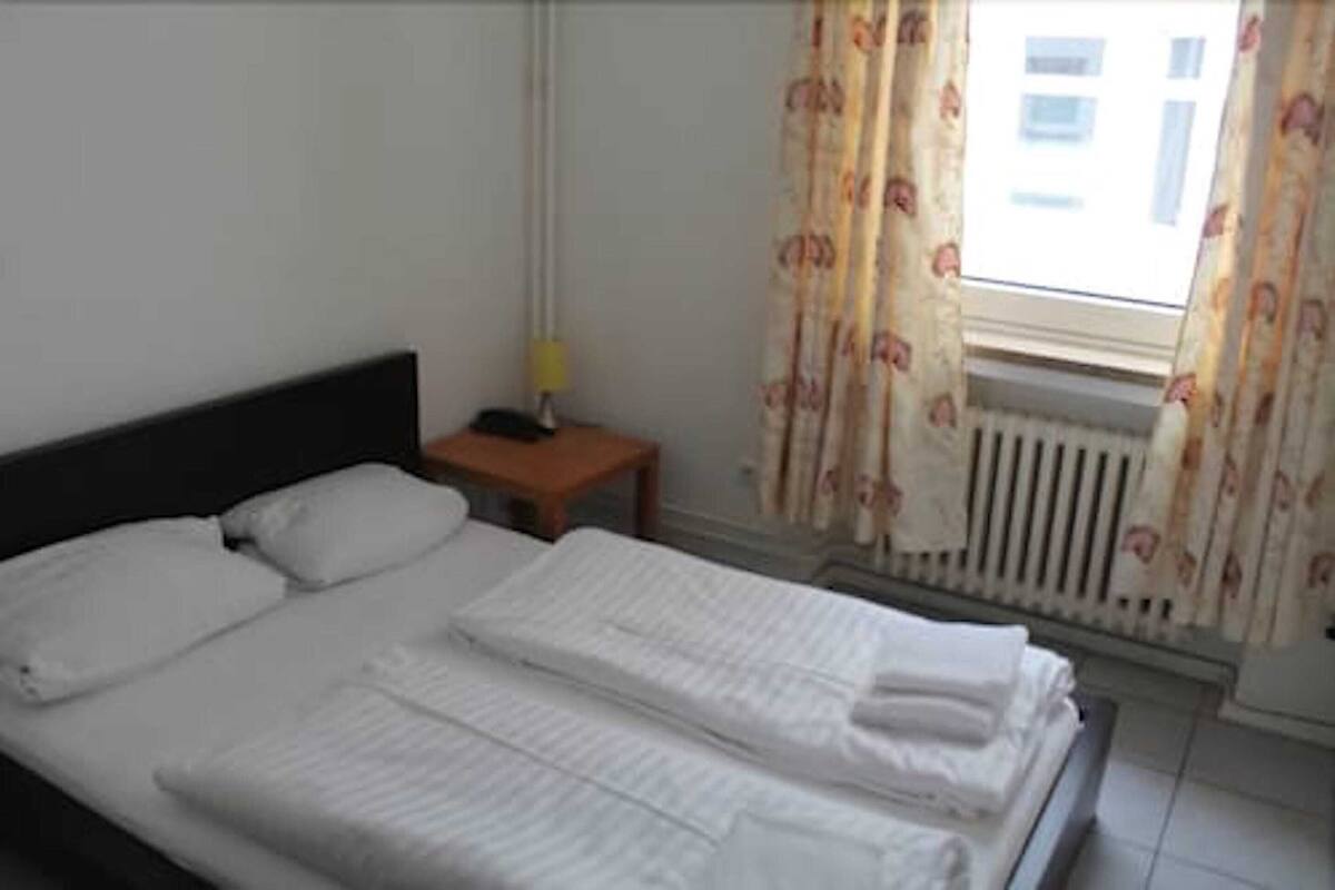 A neatly made bed with white linens, a bedside table with a lamp, and a window with floral curtains.