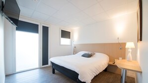Quarto de hotel com cama, mesa de cabeceira, luminária, janela e ventilador de teto.