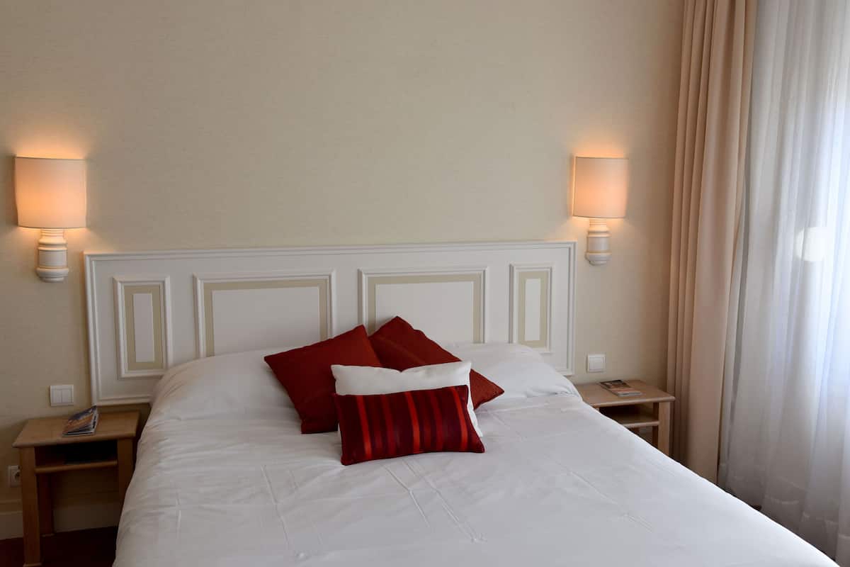 Una cama bien hecha con ropa blanca y almohadas rojas, dos mesitas de noche con lámparas, y una ventana con cortinas.