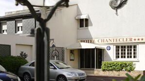 Um hotel com uma placa que diz "HOTEL CHANTECLER" e um emblema de galo. Há dois carros estacionados em frente.