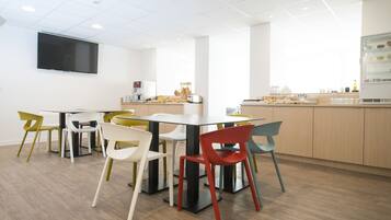 Una sala riunioni moderna con un grande tavolo e sedie colorate, una TV a schermo piatto sulla parete e un angolo cottura con elettrodomestici e generi alimentari.