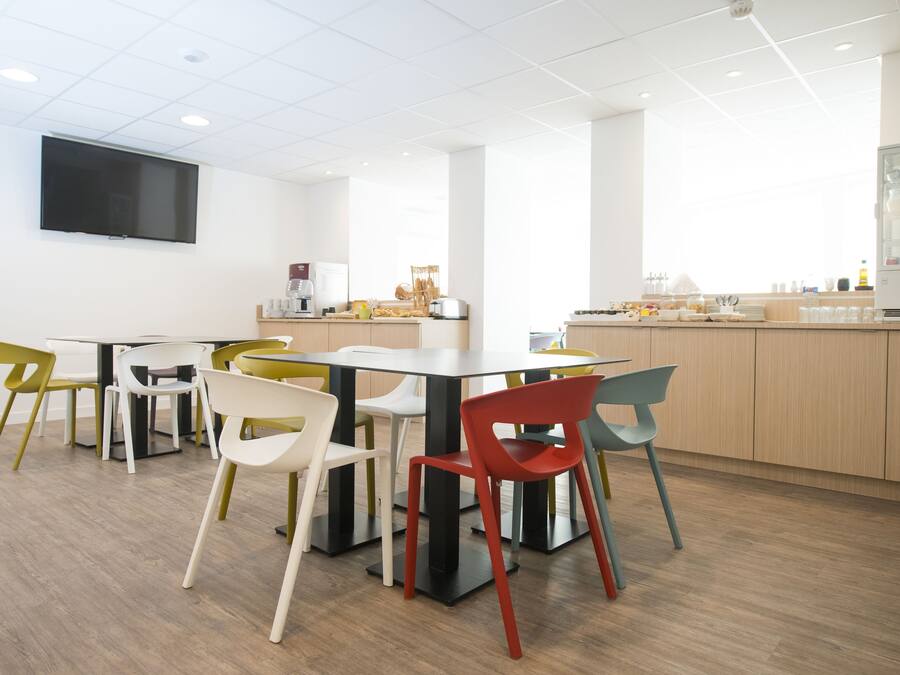 Una sala riunioni moderna con un grande tavolo e sedie colorate, una TV a schermo piatto sulla parete e un angolo cottura con elettrodomestici e generi alimentari.