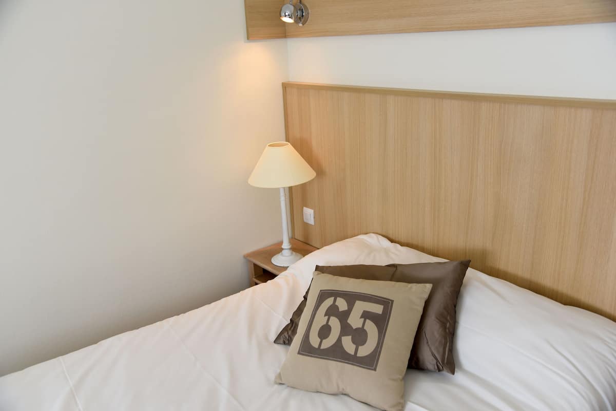 Una cama bien hecha con un cojín que lleva el número 65, una lámpara de noche y un cabecero de madera.