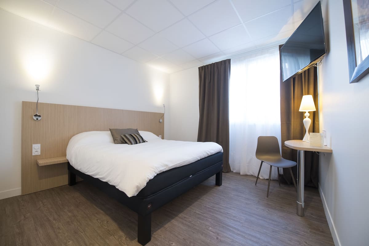 Una habitación de hotel con cama, mesita de noche, lámpara, silla y televisor en la pared.