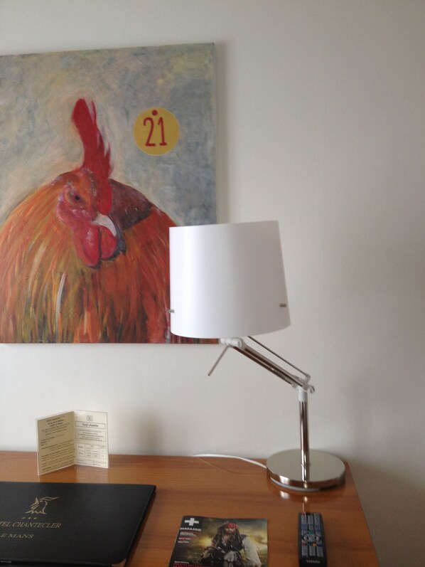A desk with a lamp, a magazine, a remote control, and a framed artwork of a rooster.