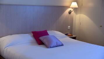 Un lit bien fait, avec des oreillers blancs et violets, une table de chevet avec une lampe, et une porte visible au fond.