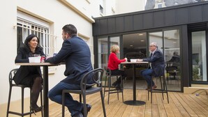 Área externa com mesas e cadeiras, piso de madeira e edifícios circundantes com grandes janelas e design moderno.