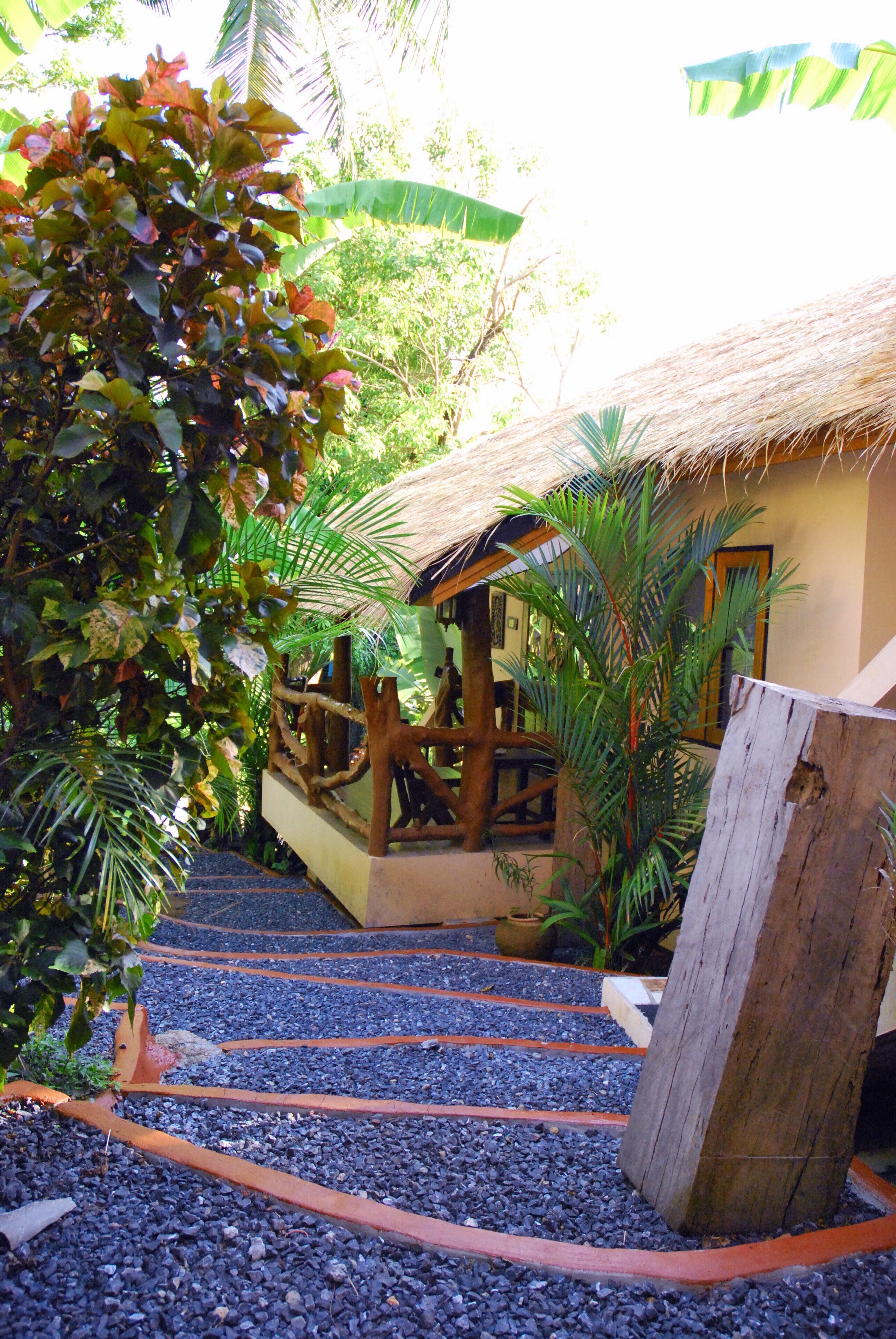 A tropical garden pathway leading to a thatched-roof structure with a wooden railing and potted plants.