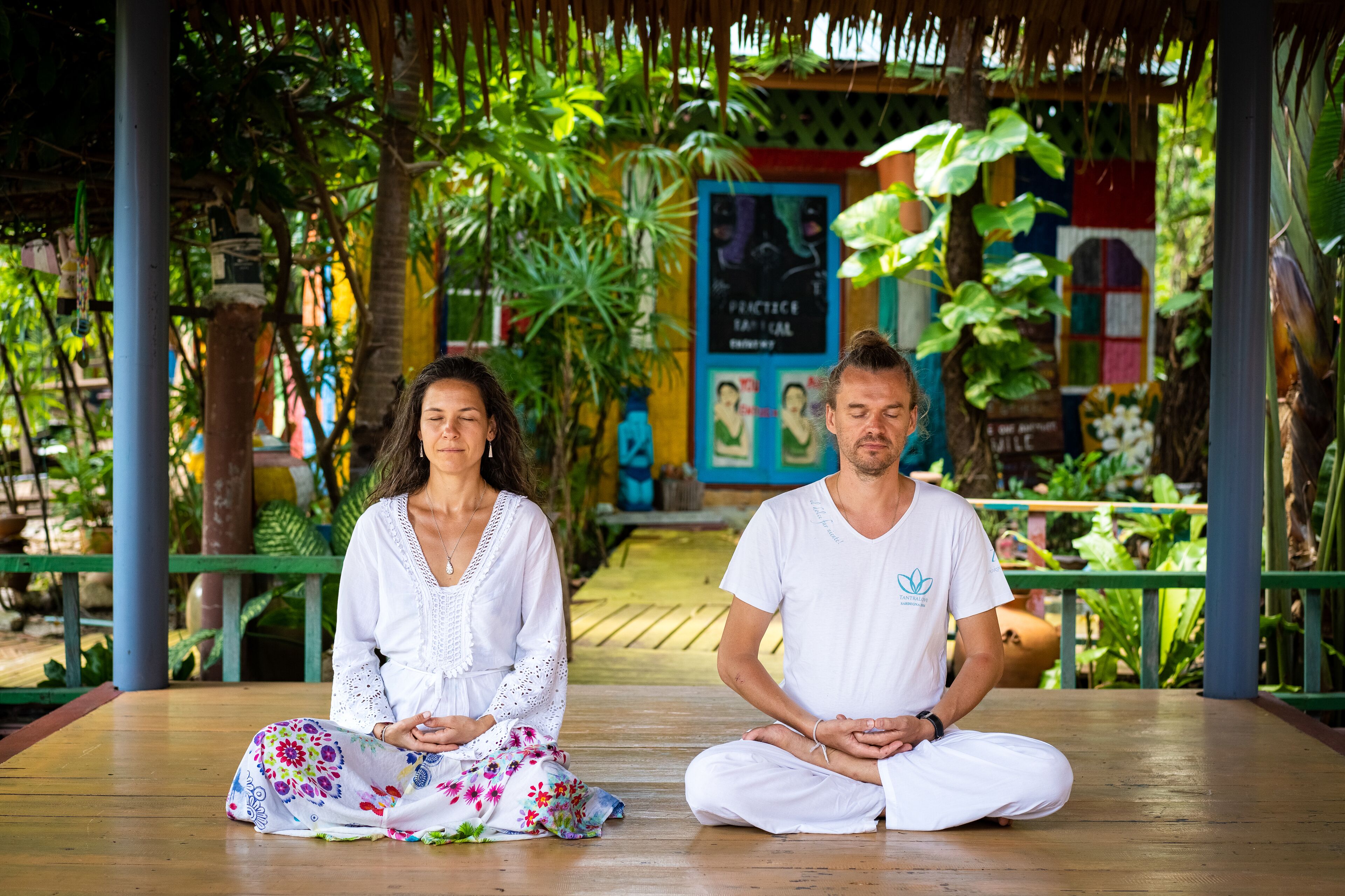 Two people meditating on a wooden platform surrounded by lush greenery and a thatched roof structure.