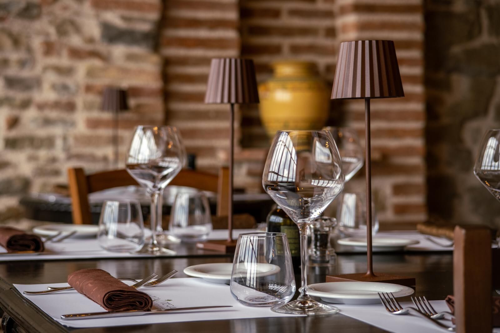 A dining table set with wine glasses, plates, and cutlery, with a brick wall and a vase in the background.