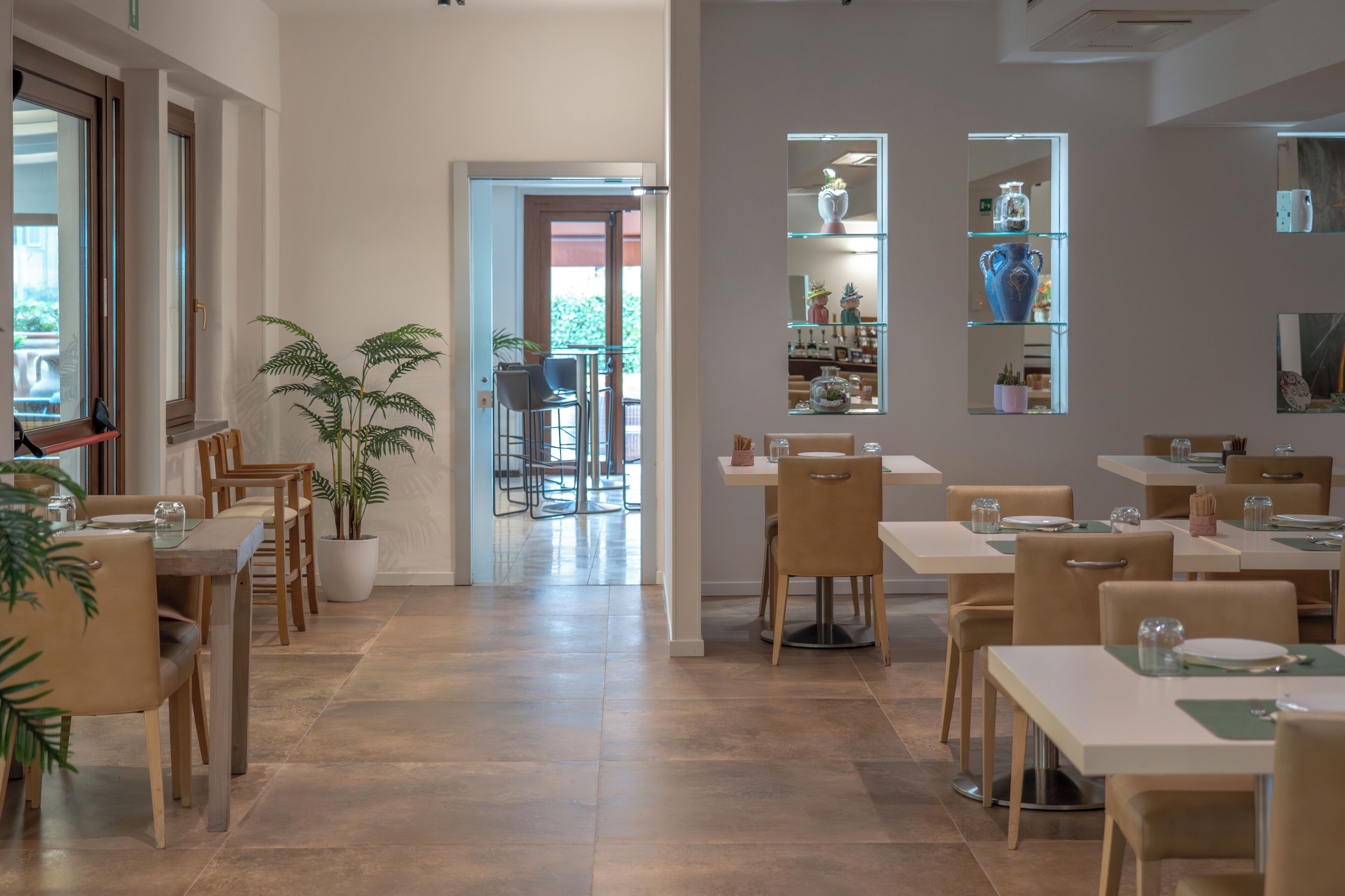 A modern dining area with multiple tables and chairs, potted plants, and a view of the outdoors.