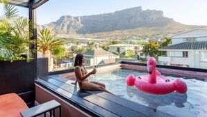 Mulher sentada à beira da piscina, com vista para as montanhas, um boi flamingo flutuante e plantas em vasos.