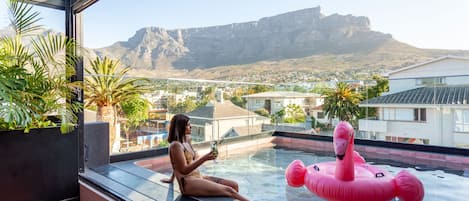 A woman sitting by a pool with a mountain view, a pink flamingo float, and potted plants.