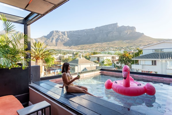 Una mujer sentada junto a una piscina con vistas a la montaña, un flotador de flamenco rosa y plantas en macetas.