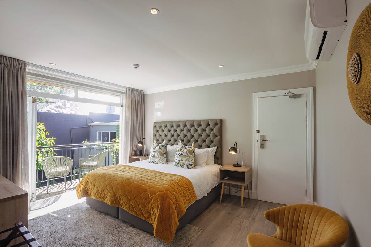 A bedroom with a large bed, a balcony with chairs, a door, and a wall-mounted air conditioner.
