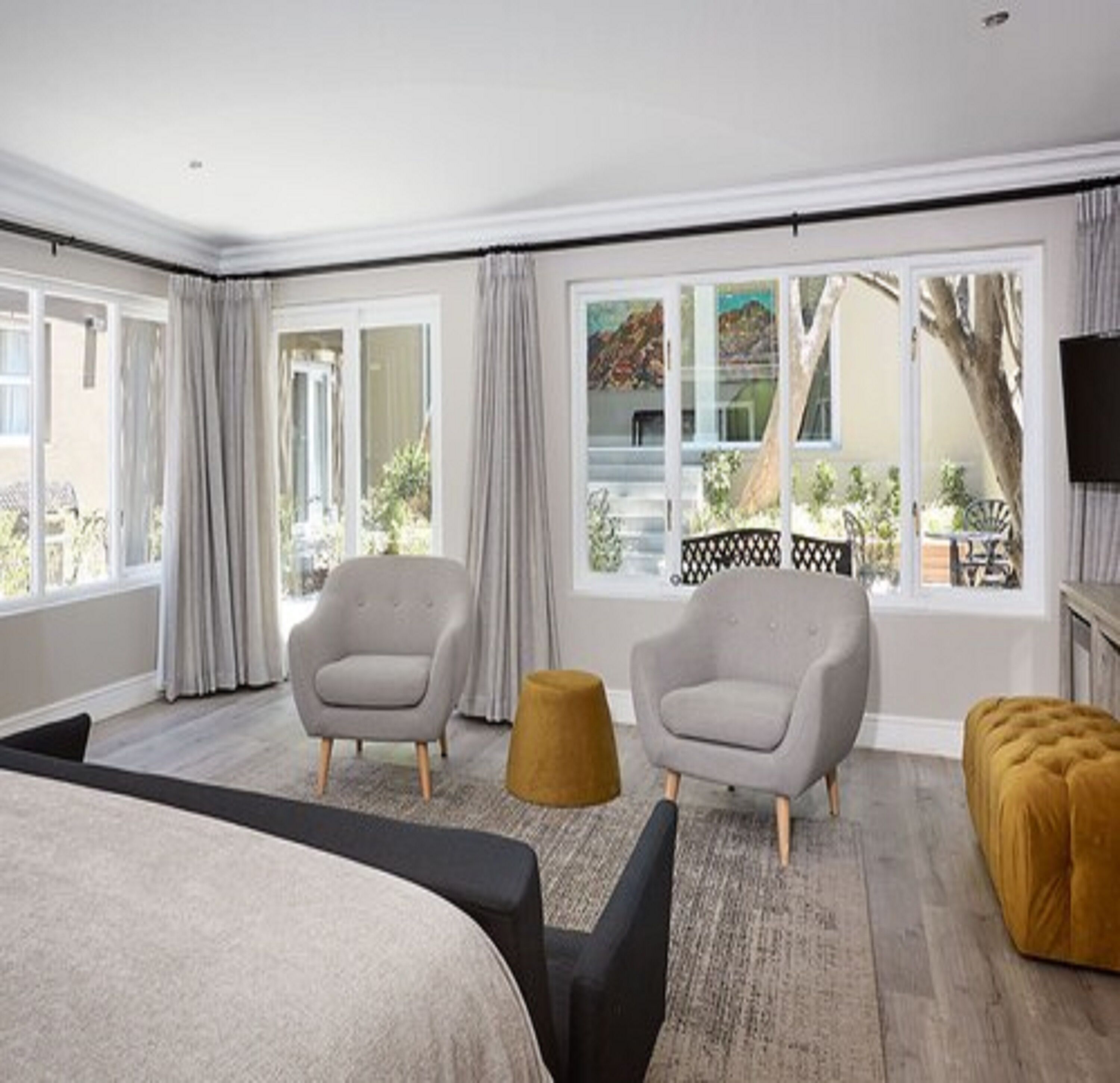A modern hotel room with a large bed, two armchairs, a small ottoman, and a view of the outdoors through large windows.