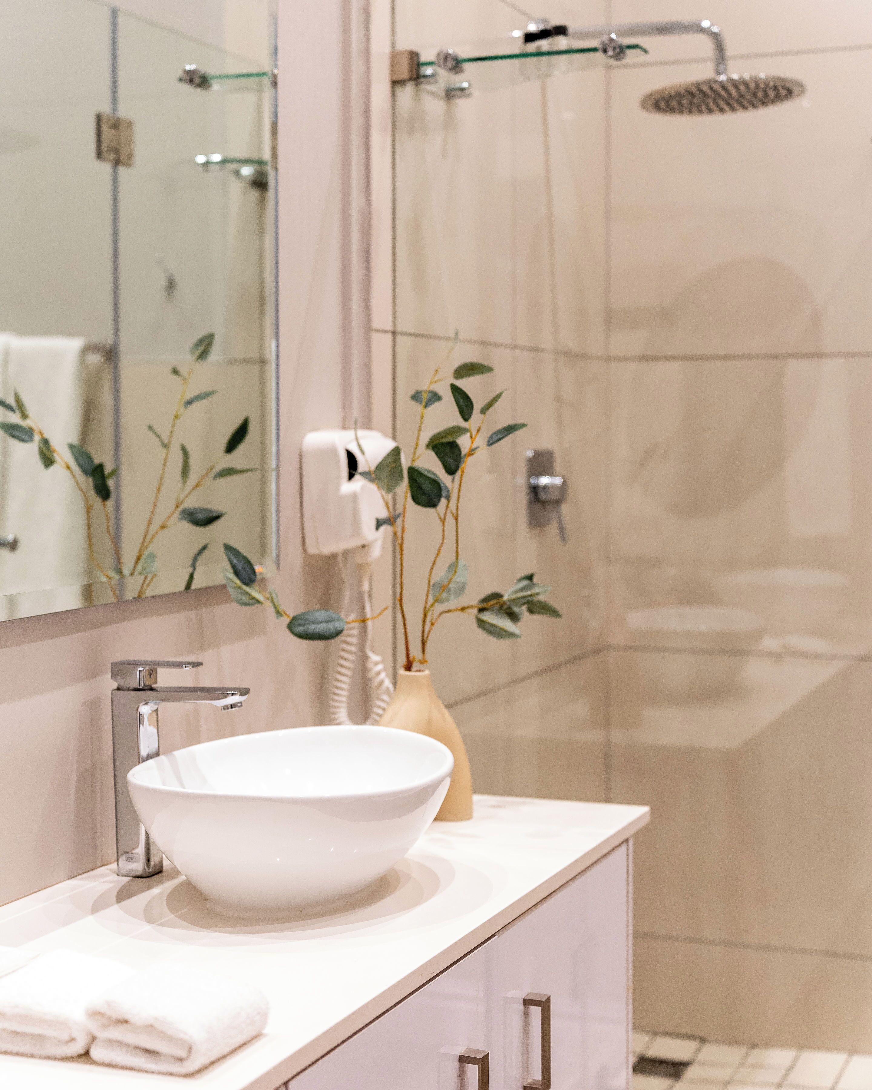 A bathroom with a white sink, a mirror, a handheld hair dryer, and a shower with a glass door.