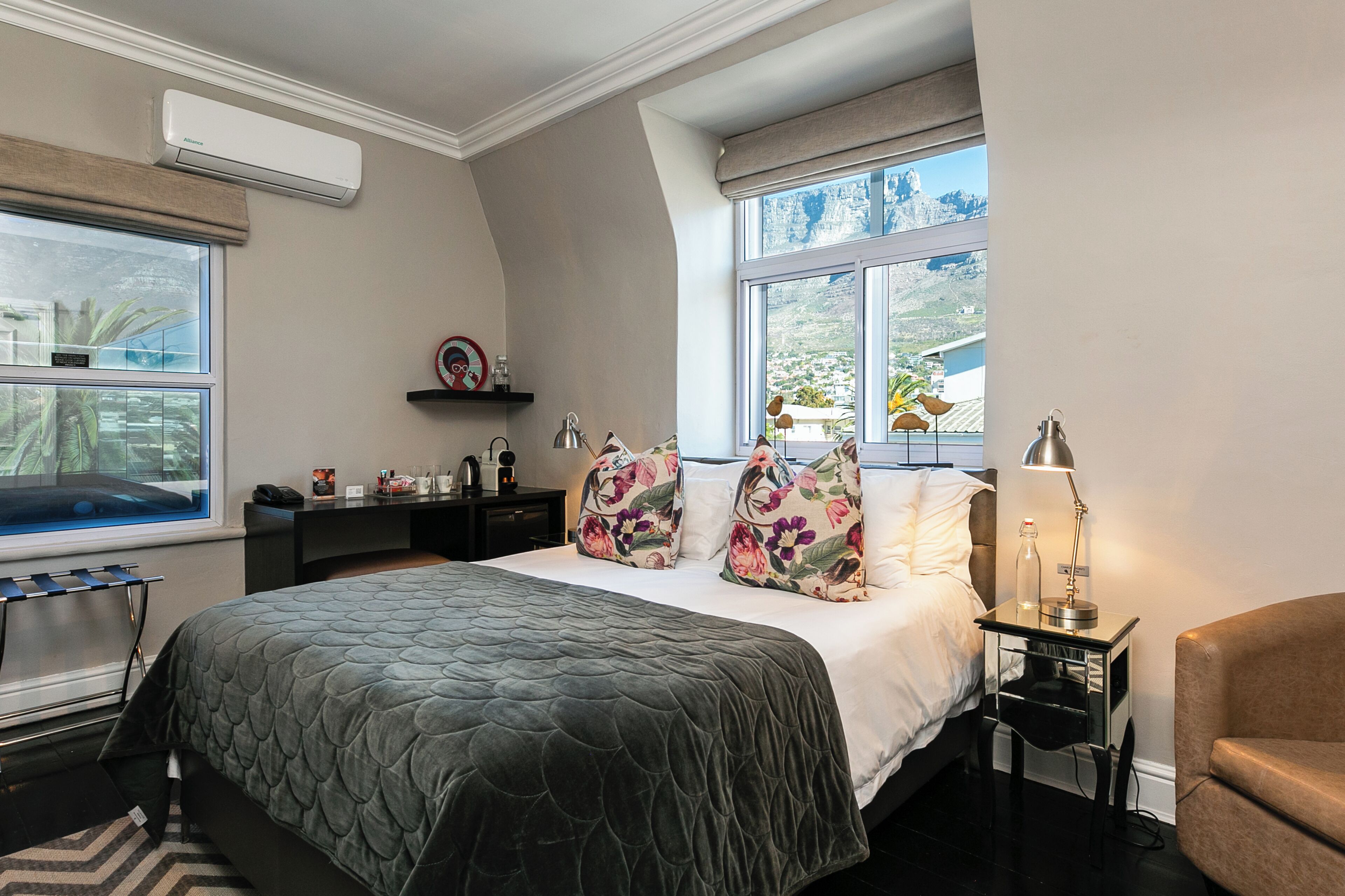 Een slaapkamer met een groot bed, een nachtkastje met een lamp, een stoel, een raam met uitzicht en een plank met decoratieve objecten.