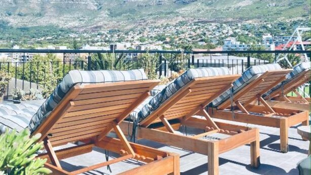 Área de lounge no terraço com espreguiçadeiras e almofadas de madeira, com vista para uma paisagem montanhosa.