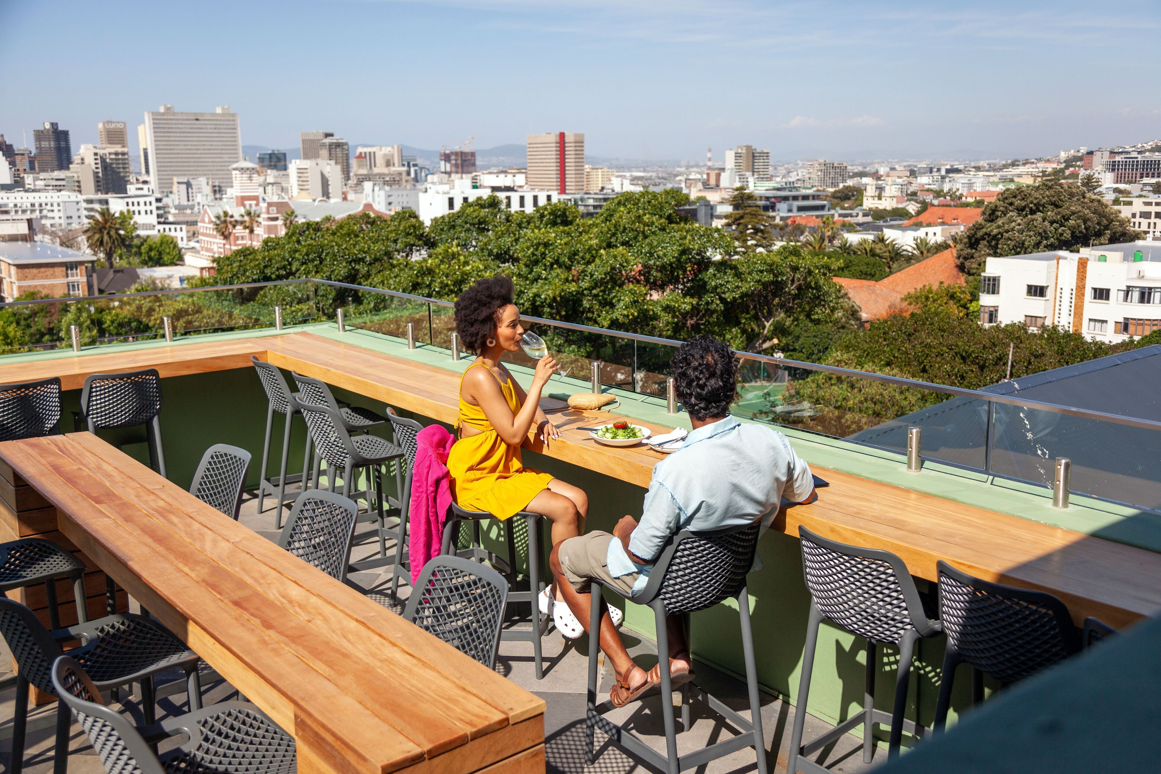An outdoor dining area with a city view, featuring a wooden table and chairs, and a glass railing.
