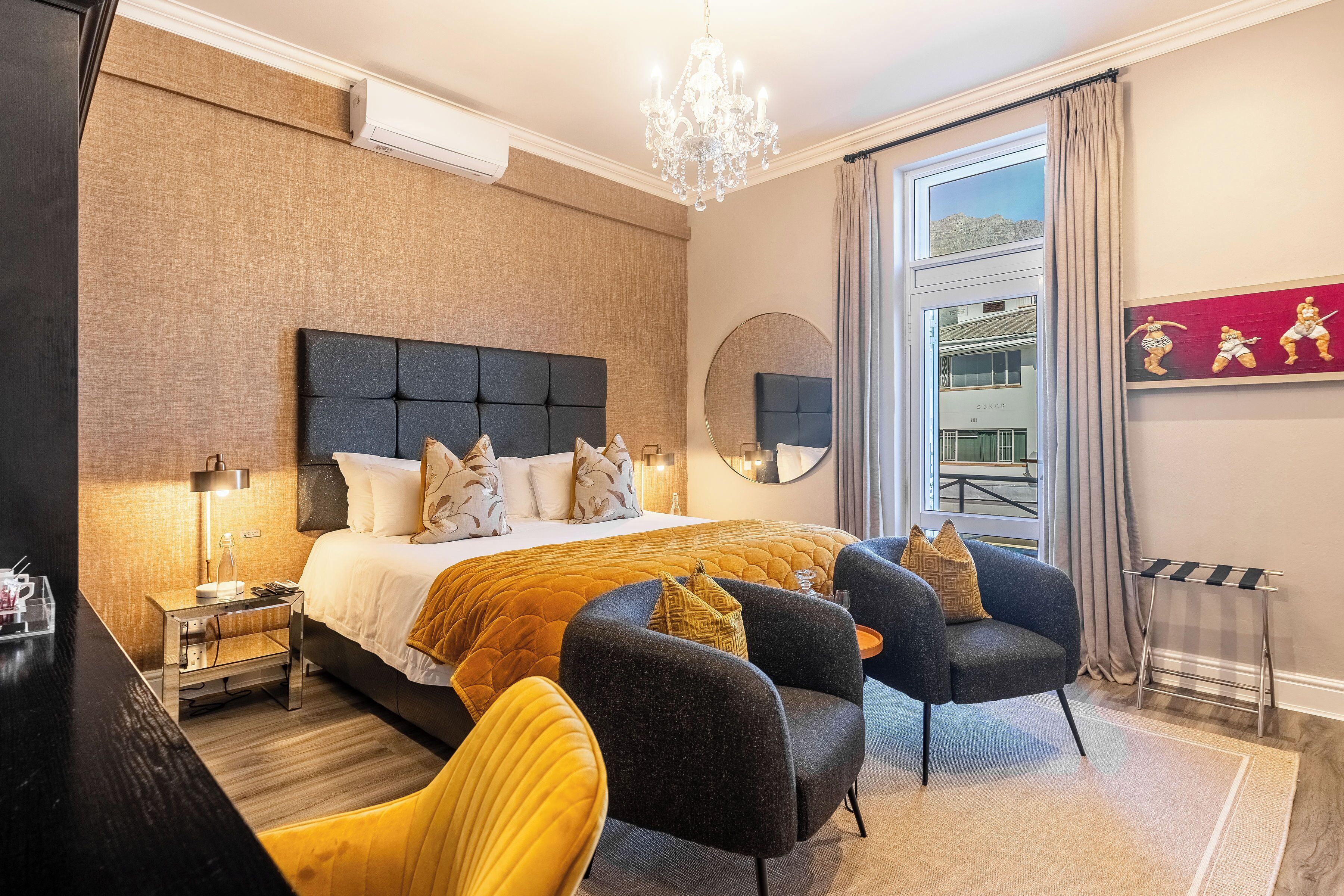 A modern hotel room with a large bed, a chandelier, a round mirror, and a view of a building through the window.