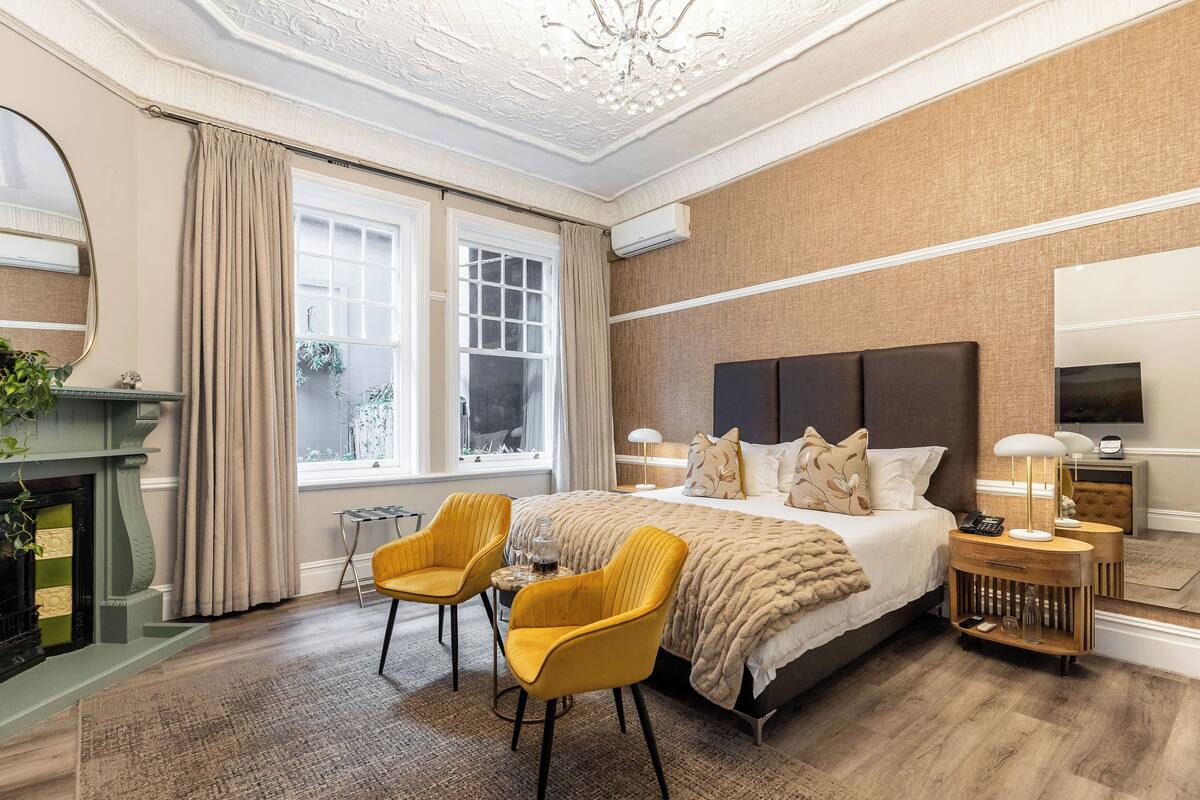 A bedroom with a large bed, two yellow chairs, a fireplace, and a large window.