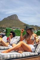 A person lounging on a striped lounge chair with a drink, a hat, and sunglasses, with a mountainous landscape in the background.