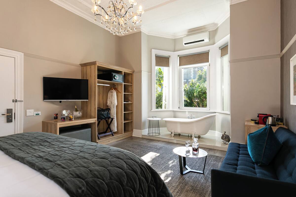 A hotel room with a bed, a TV, a wooden wardrobe, a bathroom with a bathtub, and a window with a view of greenery.