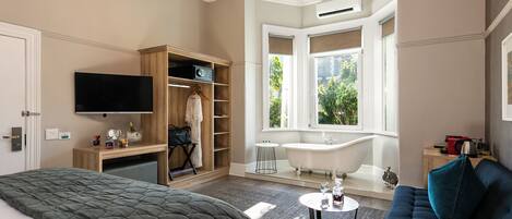 A hotel room with a bed, a TV, a wooden wardrobe, a bathroom with a bathtub, and a window with a view of greenery.