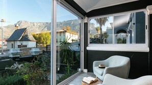 Quarto moderno com uma grande porta de vidro deslizante que dá para uma varanda, área de estar confortável com sofá e poltronas, e vista para montanhas e edifícios.